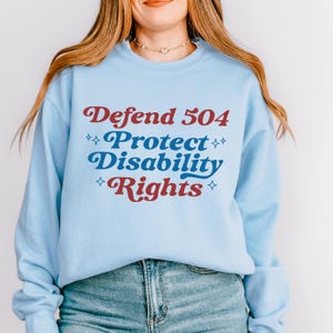 May include: A light blue sweatshirt with the text "Defend 504 Protect Disability Rights" in red and blue lettering with white stars.