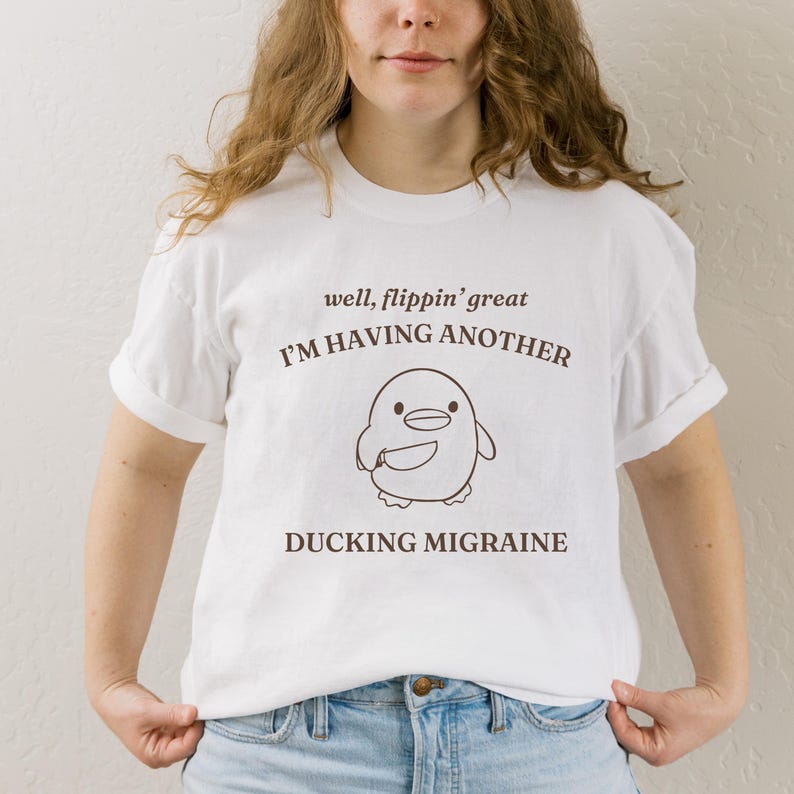May include: White t-shirt with brown text that reads "well, flippin' great I'M HAVING ANOTHER DUCKING MIGRAINE". A cartoon penguin with a knife is pictured below the text.