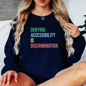 May include: A navy blue sweatshirt with the text "DENYING ACCESSIBILITY IS DISCRIMINATION" in rainbow colors.