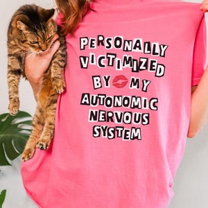 May include: Pink t-shirt with black text that reads "PERSONALLY VICTIMIZED BY MY AUTONOMIC NERVOUS SYSTEM" with a red lip print between "BY" and "MY".