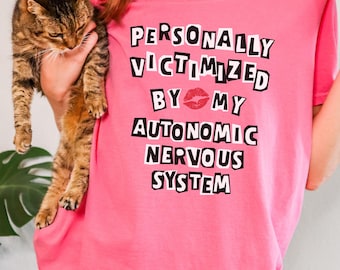 Puede incluir: Camiseta rosa con texto negro que dice "PERSONALLY VICTIMIZED BY MY AUTONOMIC NERVOUS SYSTEM" con una impresión de labios rojos entre "BY" y "MY".