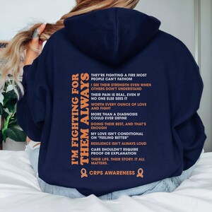Op de afbeelding: Marineblauwe hoodie met de oranje tekst "I'M FIGHTING FOR THEM ALWAYS". De achterkant toont een lijst met steunbetuigingen en de woorden "CRPS AWARENESS" met een oranje lintsymbool.