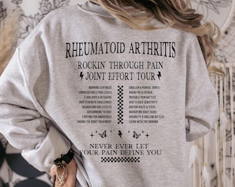 May include: Gray sweatshirt with the text "Rheumatoid Arthritis Rockin' Through Pain Joint Effort Tour" and a list of symptoms and challenges associated with the condition. The text "Never Ever Let Your Pain Define You" is printed below the list.