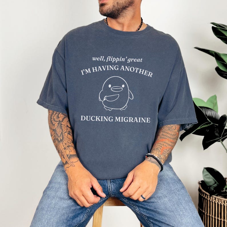 May include: A dark blue t-shirt with a white graphic of a cartoon penguin holding a knife. The text "well, flippin' great I'M HAVING ANOTHER DUCKING MIGRAINE" is printed on the shirt.