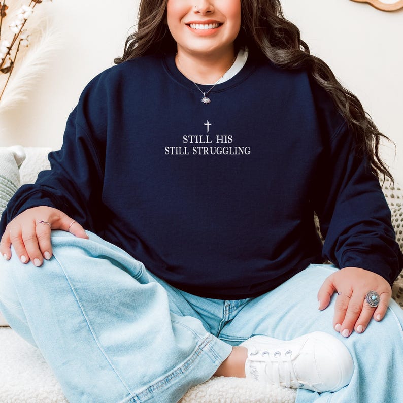 以下が含まれることがあります： 「Still His Still Struggling」というテキストと、その上に小さな十字架が書かれたネイビーブルーのスウェットシャツです。