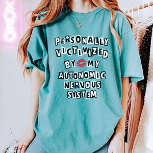 May include: A teal blue t-shirt with the text "PERSONALLY VICTIMIZED BY 💋 MY AUTONOMIC NERVOUS SYSTEM" printed in black.
