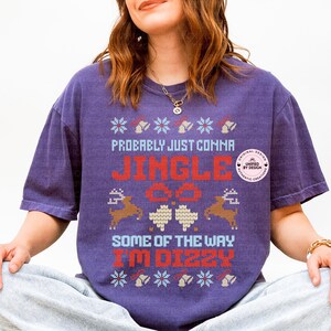Peut inclure: T-shirt violet avec un motif festif. Le t-shirt affiche le texte "Probably Just Comma Jingle All The Way I'm Dizzy" en rouge et bleu, avec des motifs de rennes et de flocons de neige. Le t-shirt a une coupe ample.