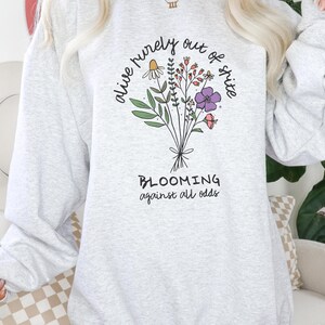 Puede incluir: Sudadera gris con un diseño floral y el texto "alive purely out of spite" y "BLOOMING against all odds".