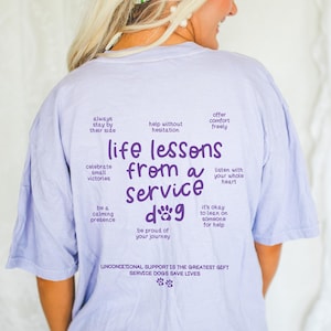 May include: Purple text on a light blue t-shirt reads "life lessons from a service dog" with a paw print graphic and a list of life lessons, including "always stay by their side", "help without hesitation", "offer comfort freely", "celebrate small victories", "be a calming presence", "listen with your whole heart", "it's okay to lean on someone for help", and "be proud of your journey". The bottom of the shirt reads "Unconditional support is the greatest gift. Service dogs save lives."