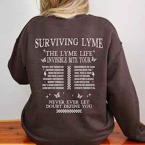 Puede incluir: Una sudadera marrón con texto blanco. El texto dice "SURVIVING LYME" y "THE LYME LIFE". Debajo hay listas de síntomas y frases relacionadas con la enfermedad de Lyme. También se incluye la frase "NEVER EVER LET DOUBT DEFINE YOU".
