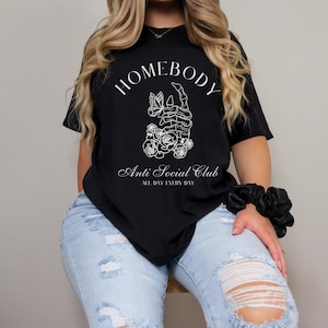 May include: Black t-shirt with white text that reads "Homebody Anti Social Club All Day Every Day". The graphic features a skeleton hand holding a butterfly with flowers.