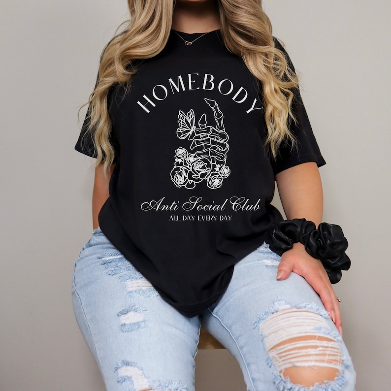 May include: Black t-shirt with white text that reads "Homebody Anti Social Club All Day Every Day". The graphic features a skeleton hand holding a butterfly with flowers.