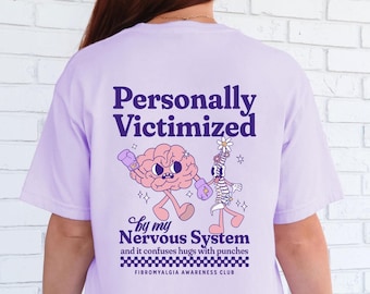 May include: A lavender t-shirt with a graphic of a brain and a skeleton boxing. The text on the shirt reads "Personally Victimized by my Nervous System and it confuses hugs with punches. Fibromyalgia Awareness Club."