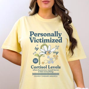 May include: Yellow t-shirt with a graphic design that says "Personally Victimized by my Cortisol Levels" with a cartoon daisy and a chemical structure. The text below the graphic says "when hormones wreak havoc it hits everything" and "Cushing's Syndrome Awareness".