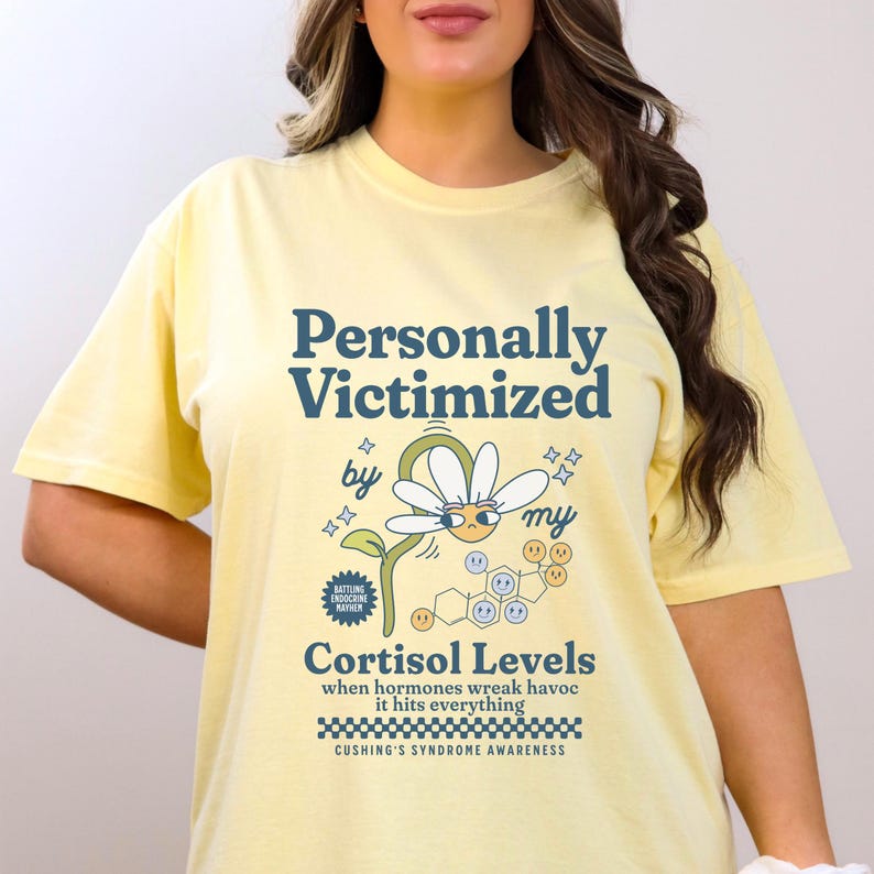May include: Yellow t-shirt with a graphic design that says "Personally Victimized by my Cortisol Levels" with a cartoon daisy and a chemical structure. The text below the graphic says "when hormones wreak havoc it hits everything" and "Cushing's Syndrome Awareness".