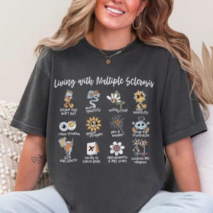 Pode incluir: Uma camiseta cinza escura com o texto "Vivendo com Esclerose Múltipla" e uma série de ilustrações de desenhos animados que representam os sintomas comuns da doença. As ilustrações incluem fadiga, espasticidade muscular, pernas instáveis, sensibilidade ao calor, problemas de visão, dificuldades de fala, picadas e agulhas, nevoeiro cerebral, efeitos colaterais, ter que cancelar planos, consultas e ressonâncias magnéticas, e recaídas de EM surpresa.