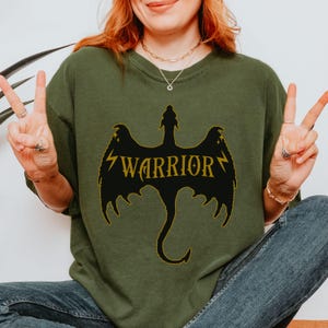 May include: An olive green sweatshirt featuring a black dragon design with the word "WARRIOR" in gold. The dragon has wings and a tail. The text is framed by lightning bolts.