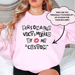 May include: Pink sweatshirt with the text "PERSONALLY VICTIMIZED BY MY CUSTOM" in black with a red lip print. The text "WHAT ARE YOU PERSONALLY VICTIMIZED BY? LET US KNOW FOR YOUR NEW SHIRT!" is in a speech bubble.