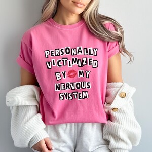 Könnte beinhalten: Pinkes T-Shirt mit dem Text "PERSONALLY VICTIMIZED BY MY NERVOUS SYSTEM" in schwarzen Buchstaben. Ein roter Lippenstift-Druck befindet sich zwischen "BY" und "MY".