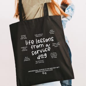 以下が含まれることがあります： 「サービスドッグからの人生の教訓」と書かれた白い文字が入った黒いトートバッグ。バッグには「いつもそばにいて」や「自分の旅を誇りに思う」などのフレーズが含まれています。バッグの底には「無条件のサポートが最大の贈り物」と書かれています。