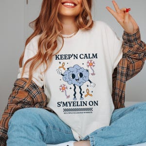 Puede incluir: Una camiseta blanca con el texto "Keep'n Calm S'Myelin On" y un cerebro de dibujos animados con cintas naranjas. El texto "Multiple Sclerosis Warrior" está debajo del cerebro.
