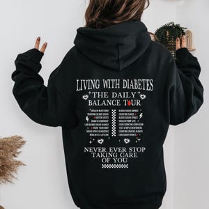 K&ouml;nnte beinhalten: Schwarzer Hoodie mit einem wei&szlig;en Grafikdruck, der "Leben mit Diabetes - Die t&auml;gliche Balance Tour" sagt und die Herausforderungen des Lebens mit Diabetes auflistet. Die Grafik enth&auml;lt auch zwei rote Herzen und den Text "H&ouml;r niemals auf, auf dich aufzupassen".