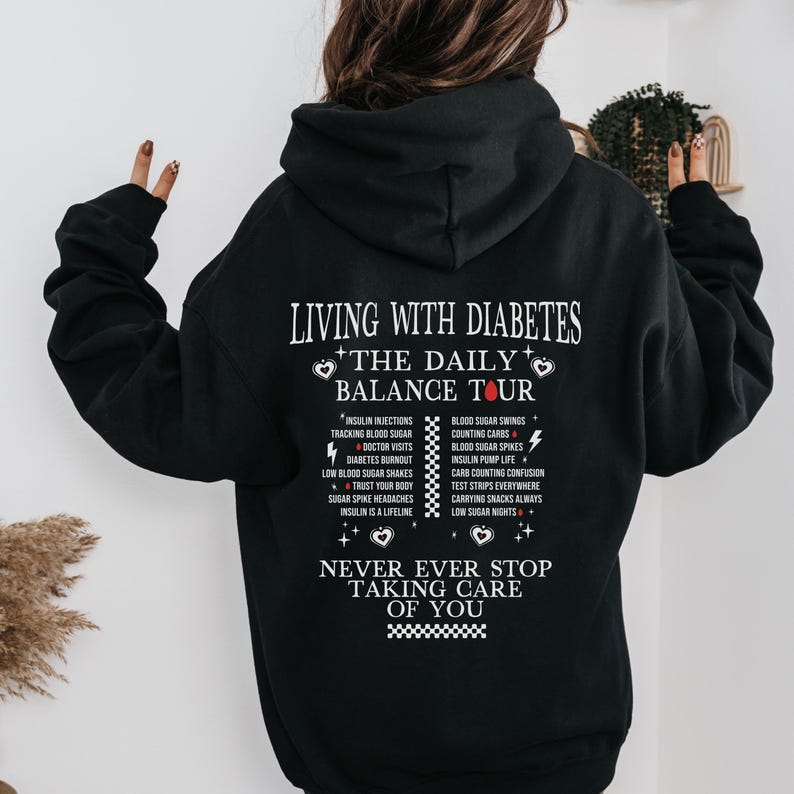 K&ouml;nnte beinhalten: Schwarzer Hoodie mit einem wei&szlig;en Grafikdruck, der "Leben mit Diabetes - Die t&auml;gliche Balance Tour" sagt und die Herausforderungen des Lebens mit Diabetes auflistet. Die Grafik enth&auml;lt auch zwei rote Herzen und den Text "H&ouml;r niemals auf, auf dich aufzupassen".