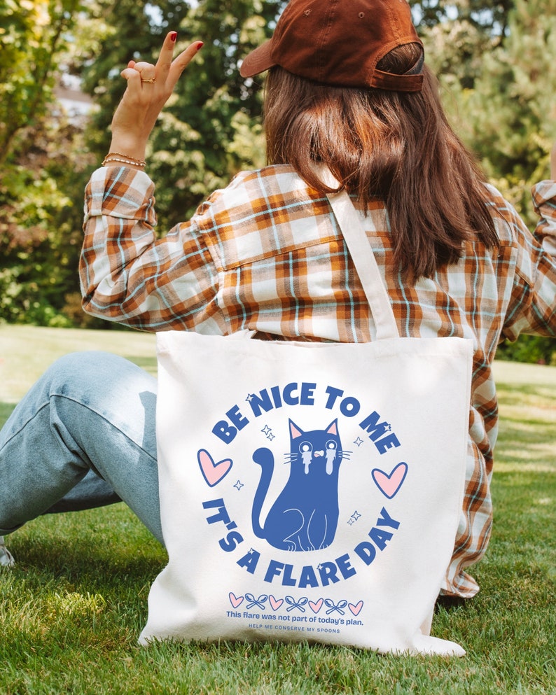 May include: White canvas tote bag featuring a blue cat illustration and the text "Be Nice To Me It's A Flare Day". The text is in a pink heart with a blue outline. The cat has blue tears and is sitting with its tail curled around its legs. The text "This flare was not part of today's plan. Help me conserve my spoons" is printed below the illustration.