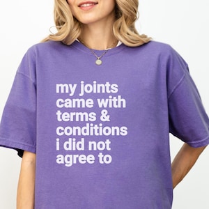 Könnte beinhalten: Ein lilafarbenes Kurzarm-T-Shirt mit dem weißen Text "my joints came with terms & conditions i did not agree to". Das Shirt ist einfarbig und hat eine lockere Passform.
