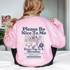 Puede incluir: Sudadera rosa con un gráfico de dos pulmones con mascarillas y el texto "Please Be Nice To Me" y "Every Day Is A Marathon but I'm here and I'm being really BRAVE about it LONG COVID AWARENESS CLUB".