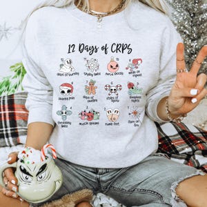 Op de afbeelding: Een grijs sweatshirt met een witte grafische print van de "12 dagen van CRPS" met een kerstthema. De grafiek toont 12 verschillende illustraties die symptomen van CRPS voorstellen, zoals branden, tintelen, zenuwstoornissen en slapeloze nachten.
