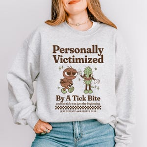 Puede incluir: Una sudadera gris claro con el texto "Personally Victimized By A Tick Bite" y un gráfico de dibujos animados de una hormiga y un pepinillo. También se incluye el texto "LYME DISEASE AWARENESS CLUB".
