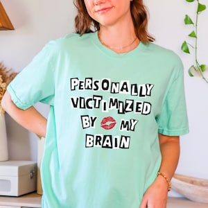 May include: A light green t-shirt with the text "PERSONALLY VICTIMIZED BY MY BRAIN" in black block letters with a red lip print between the words "BY" and "MY".