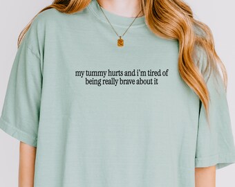 Puede incluir: Camiseta verde claro con el texto negro "my tummy hurts and I'm tired of being really brave about it". La camiseta tiene mangas cortas y cuello redondo.