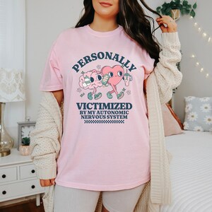K&ouml;nnte beinhalten: Pinkes T-Shirt mit einem Grafikdruck eines Gehirns und eines Herzens, die Getr&auml;nke halten. Der Text lautet "PERSONALLY VICTIMIZED BY MY AUTONOMIC NERVOUS SYSTEM".