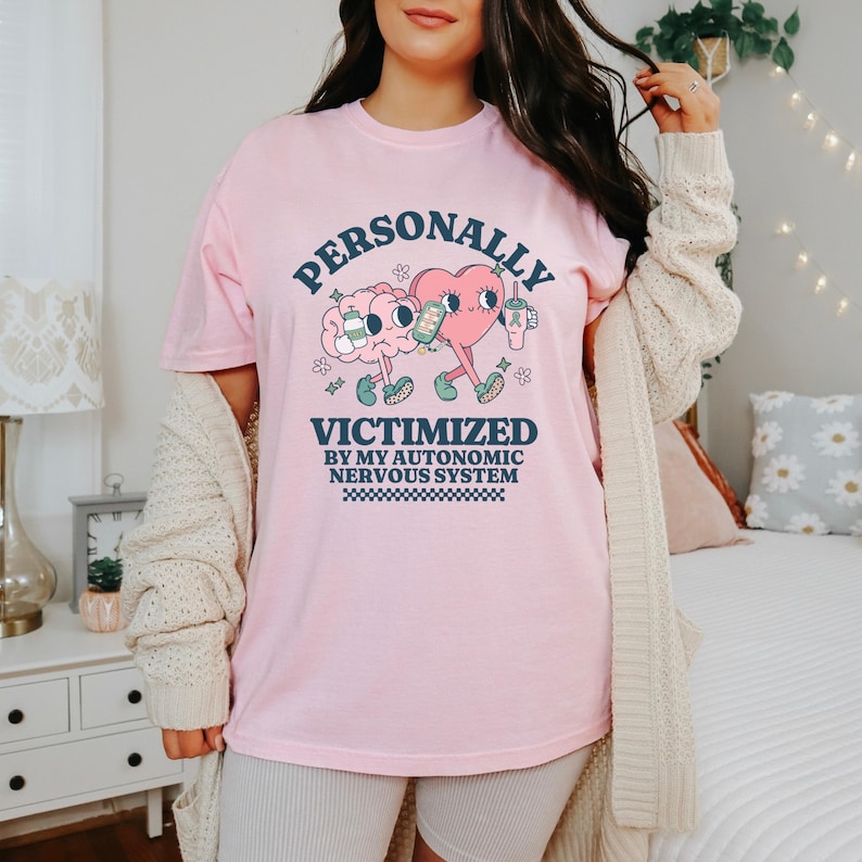 K&ouml;nnte beinhalten: Pinkes T-Shirt mit einem Grafikdruck eines Gehirns und eines Herzens, die Getr&auml;nke halten. Der Text lautet "PERSONALLY VICTIMIZED BY MY AUTONOMIC NERVOUS SYSTEM".