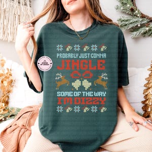Peut inclure: T-shirt vert fonc&eacute; avec un motif festif. Le t-shirt pr&eacute;sente le texte "Probably Just Comma Jingle All The Way I'm Dizzy" en rouge et blanc, ainsi que des flocons de neige, des cloches, des rennes et des tr&egrave;fles. Un autocollant rond est visible.