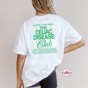 Op de afbeelding: Wit T-shirt met groene tekst: "GLUTEN. IS. NOT. SAFE. THE CELIAC DISEASE Club." Het shirt bevat ook een beschrijving van coeliakie. Onderdeel van de collectie "UNIFIED BY DESIGN AUTHENTIC CREATIONS".