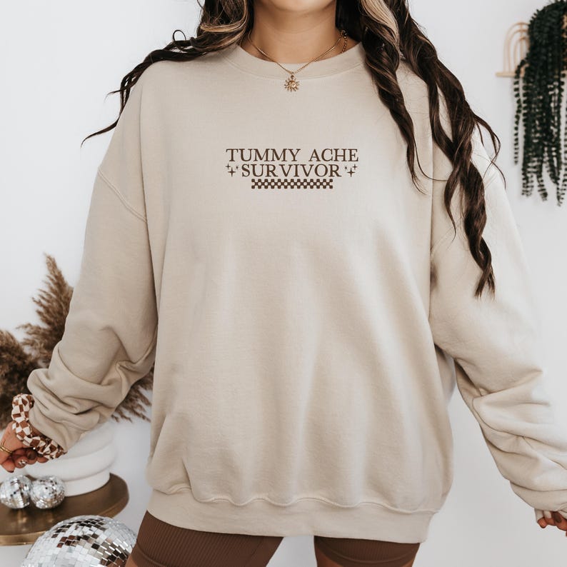 May include: Beige sweatshirt with the text "TUMMY ACHE SURVIVOR" in brown lettering. The text is above a checkered pattern. The sweatshirt has a crew neck and long sleeves.