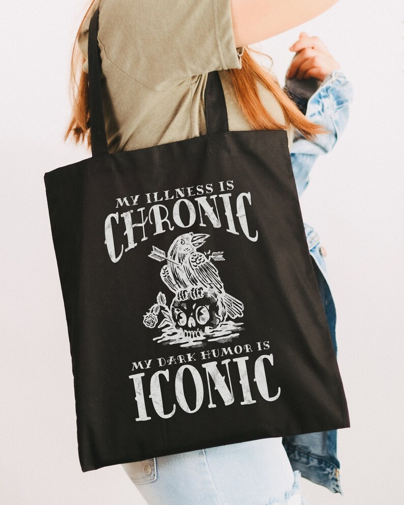 Black canvas tote bag with a goth inspired design that includes the phrase, "my illness is chronic, my dark humor is iconic"