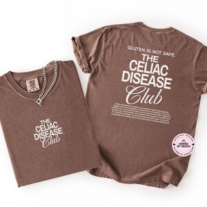Op de afbeelding: Twee bruine t-shirts met witte tekst met de tekst "THE CELIAC DISEASE Club". E&eacute;n shirt heeft een zilveren ketting. De achterkant van &eacute;&eacute;n shirt luidt "GLUTEN. IS. NOT. SAFE." en heeft een beschrijving van Coeliakie.
