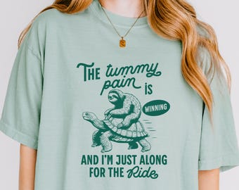 Puede incluir: Camiseta verde claro con el texto "The tummy pain is winning" y "And I'm just along for the ride." El gráfico muestra un perezoso montando una tortuga. La camiseta es de un material suave.