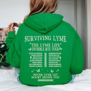 Puede incluir: Sudadera con capucha verde con texto blanco que dice "Surviving Lyme - The Lyme Life - Invisible Bite Tour". El texto enumera los síntomas y los desafíos asociados con la enfermedad de Lyme. La parte inferior de la sudadera dice "Never ever let doubt define you".