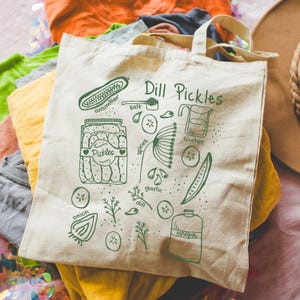 May include: A white canvas tote bag with a green illustration of ingredients for dill pickles. The text "Dill Pickles" is written in green at the top of the bag. The ingredients include cucumber, salt, spices, garlic, dill, onion, water, and vinegar.