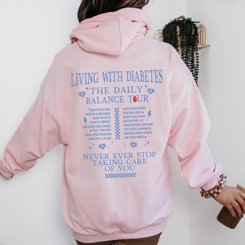 K&ouml;nnte beinhalten: Rosafarbener Hoodie mit einem blauen und roten Grafikdruck, der "Leben mit Diabetes + Die t&auml;gliche Balance-Tour" sagt und die t&auml;glichen Herausforderungen des Lebens mit Diabetes auflistet. Die Grafik enth&auml;lt auch den Text "H&ouml;r niemals auf, auf dich aufzupassen".