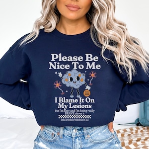 以下が含まれることがあります： 「Please Be Nice To Me」と「I Blame It On My Lesions but I'm here and I'm being really BRAVE about it STILL S'MYELIN THROUGH IT ALL」というテキストと、漫画の脳のグラフィックが描かれたネイビーブルーのスウェットシャツ。