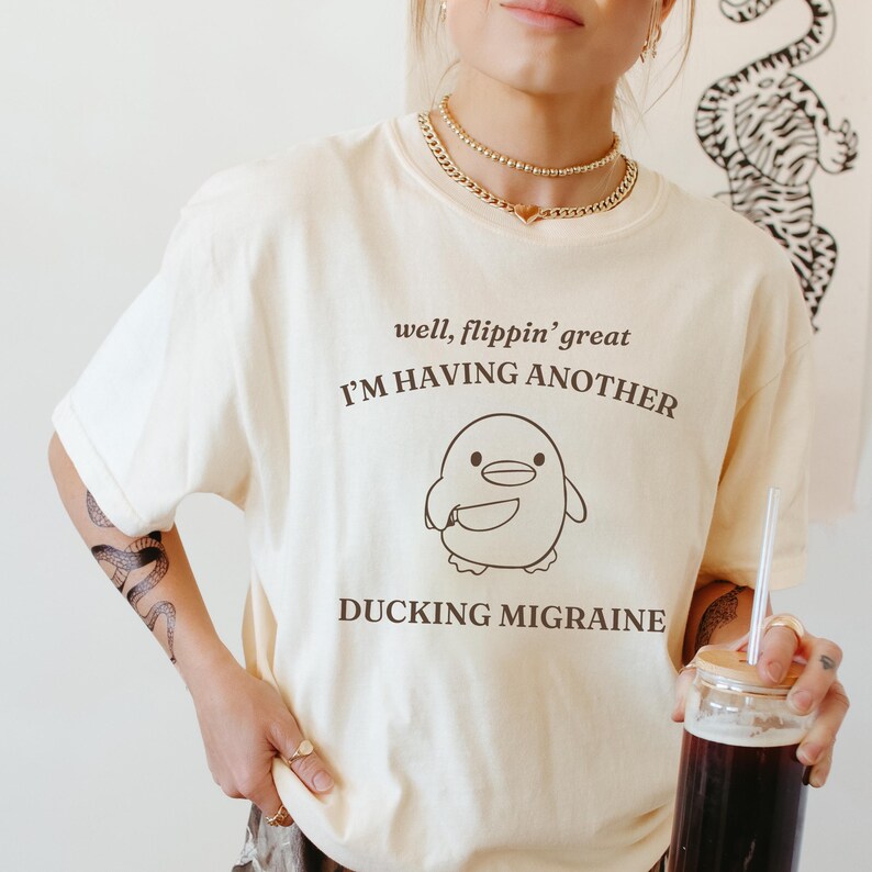 May include: A cream colored t-shirt with a cartoon penguin holding a knife and the text "well, flippin' great I'M HAVING ANOTHER DUCKING MIGRAINE".