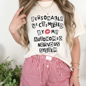 May include: A white t-shirt with the text "PERSONALLY VICTIMIZED BY MY AUTONOMIC NERVOUS SYSTEM" in black block letters. A red lip print is between the words "BY" and "MY". The person is wearing red and white gingham shorts.