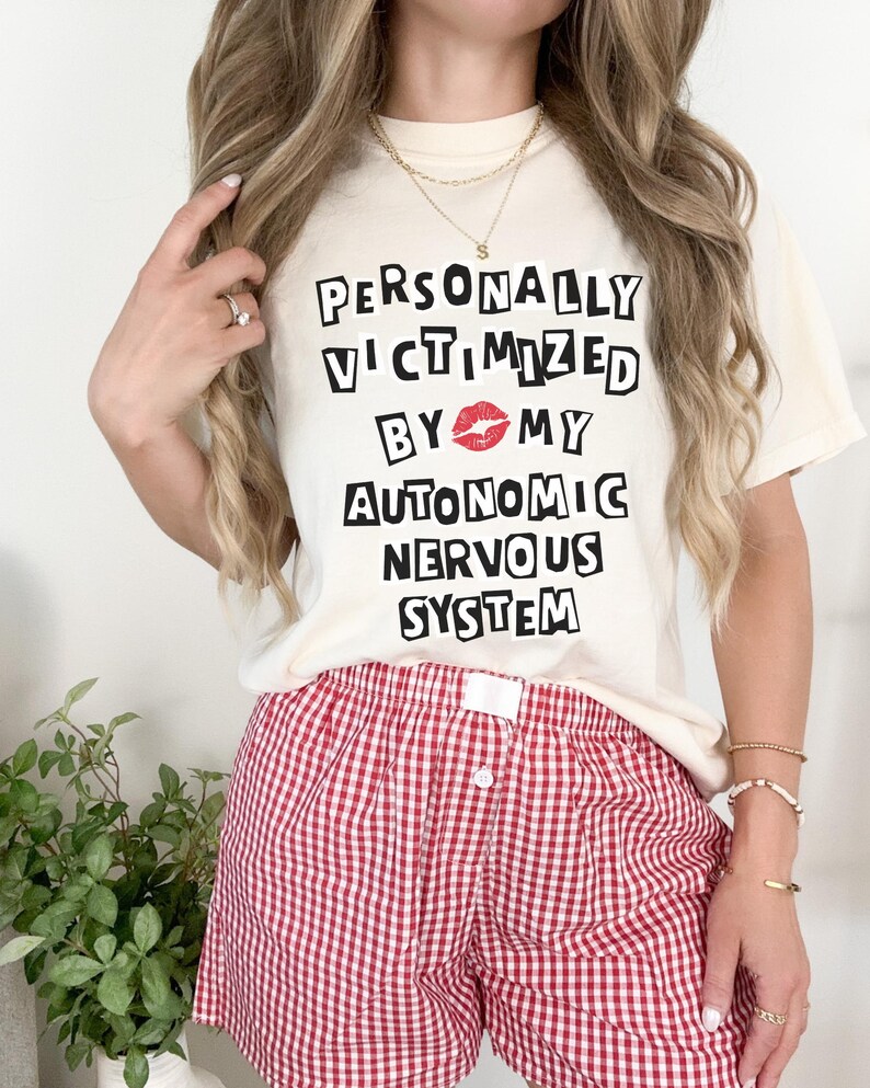 May include: A white t-shirt with the text "PERSONALLY VICTIMIZED BY MY AUTONOMIC NERVOUS SYSTEM" in black block letters. A red lip print is between the words "BY" and "MY". The person is wearing red and white gingham shorts.