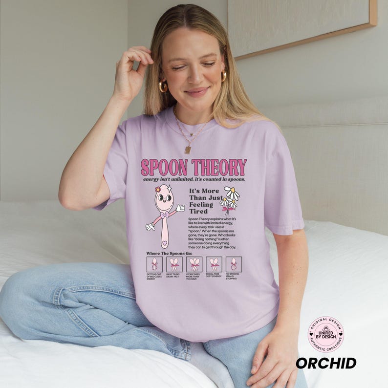 May include: A light purple t-shirt with the text "SPOON THEORY" in pink. The shirt features a cartoon spoon character and the phrase "It's More Than Just Feeling Tired." The shirt also has a graphic explaining the Spoon Theory.